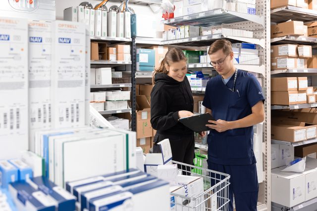 Kaksi logistikkoa tutkii yhdessä lähestyslistaa tai tilausta. He ovat sairaalan varastotilassa, joka on valoisa ja seiniä täyttävät hyllyköt. Hyllyissä on paljon erilaisia tuotteista eri kokoisissa laatikoissaan.