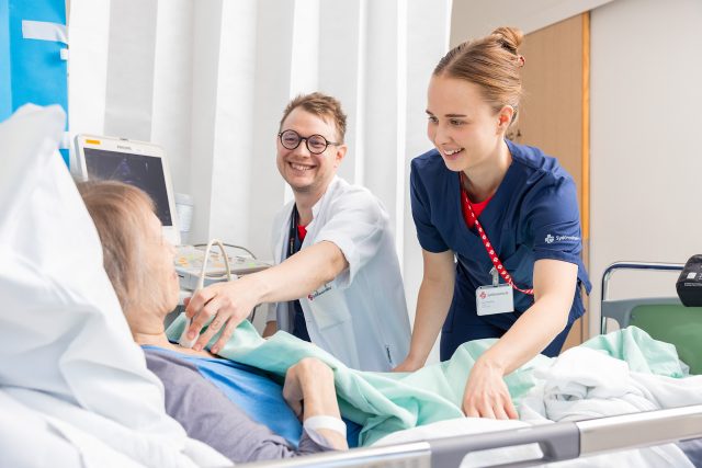 Vuodeosastolla lääkäri ja hoitaja juttelevat hymyillen potilaan kanssa, joka makaa vuoteessa. Potilasta ei näytetä.