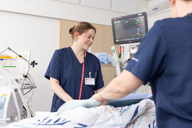 Sydänteho-osaston potilashuoneessa kaksi hoitajaa peittelee potilasta. Toinen hoitajista hymyilee potilaalle ja toinen on selin kameraan. Potilasta ei näy kuvassa. Hoitajan taustalla näkyy hoidossa käytettävää välineistöä.