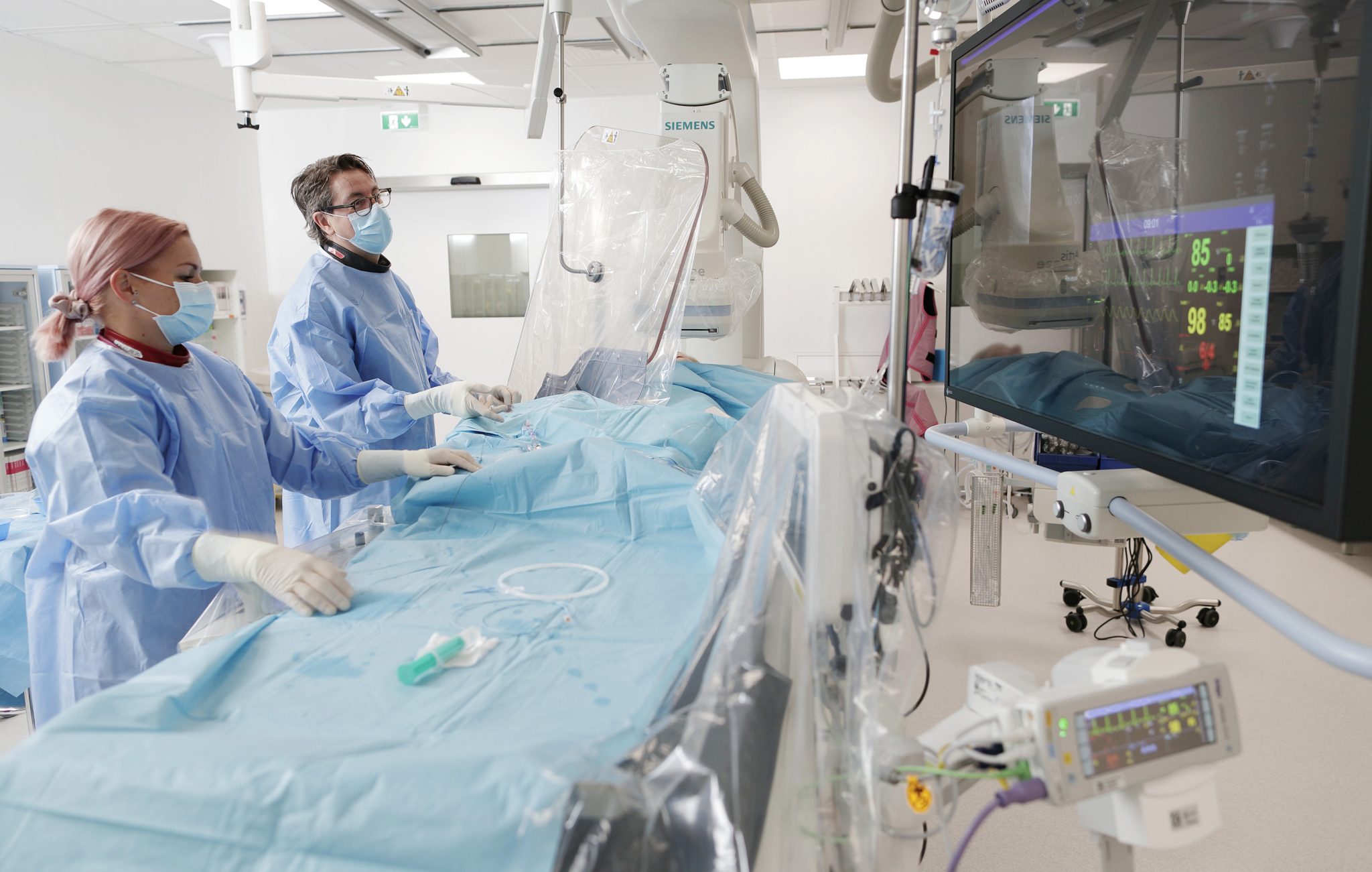 Suojavaatteisiin pukeutuneet sairaanhoitaja ja kardiologi valmistautuvat sydänpotilaan sepelvaltimoiden varjoainekuvaukseen Sydänsairaala Novan toimenpidesalissa.