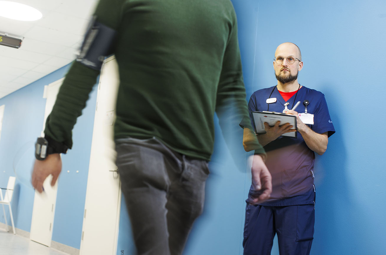 Fysioterapeutti seuraa potilaan kävelytestisuoritusta sairaalan käytävällä. Fysioterapeutilla on kädessään muistiinpanopaperit, joihin hän kirjaa tietoja samalla kun katsoo potilasta kävelemässä. Potilaalla on yllään tavalliset arkivaatteet eikä hänen kasvojaan näytetä.